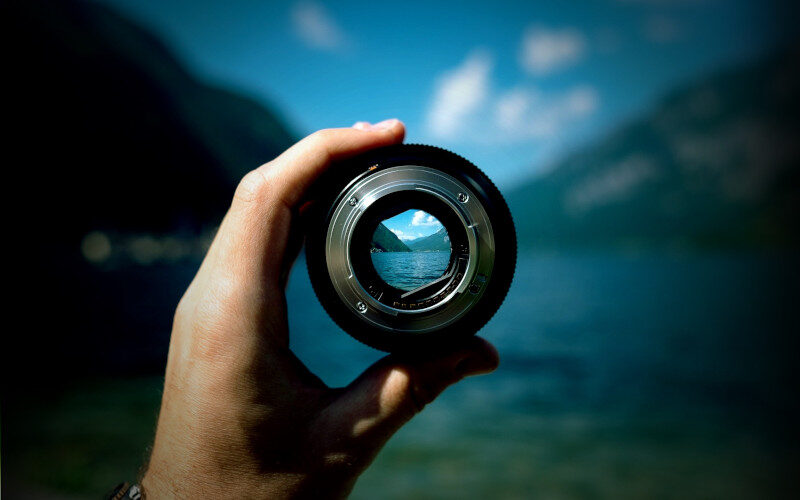 una mano tiene in mano un obiettivo fotografico che mette a fuoco un dettaglio di un paesaggio di lago sfocato