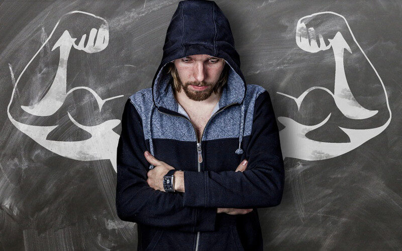 ragazzo incappucciato con aria pensierosa con alle spalle una lavagna su cui sono disegnate due braccia forzute che mostrano i muscoli